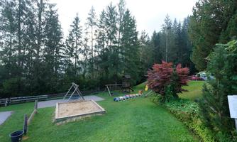 A playground with a sandbox and swings in a green environment.  
Surrounded by trees and a well-kept lawn.