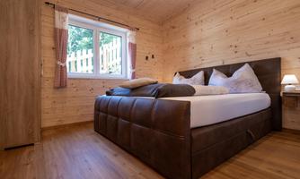 A cozy bedroom with a large double bed and wooden walls. The room has a window overlooking greenery and a warm wooden floor.
