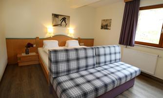 A cozy bedroom with a double bed and a gray pillow sofa. Large windows provide plenty of daylight and a pleasant atmosphere.