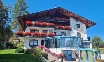 A picturesque building with blooming balconies and a large glass porch. Surrounded by green meadows and trees under a clear blue sky.