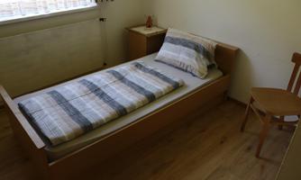 A simple bedroom with a single bed and a chair. The walls are light and the floor is wooden.