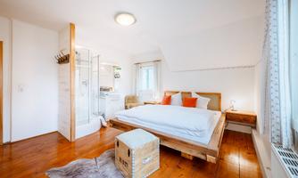 A modern bedroom with wooden flooring and a comfortable bed. The room also features a bright shower area.
