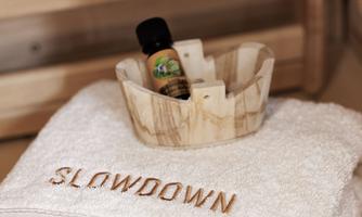 A white towel with the inscription "SLOWDOWN" is lying on a wooden surface. Additionally, there is a small bowl with a fragrance oil on it.