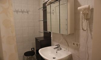 A small bathroom with a sink, a mirror, and a towel rack. The walls are covered in light tiles and there is a metal stool.