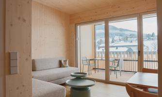 A modern living room with bright wooden paneling. There is a sofa, a table, and a balcony overlooking the snowy landscape.