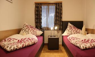 A cozy bedroom with two beds and colorful bedcovers. In front of the window stands a nightstand with a lamp.