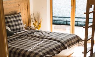 A bright bedroom with a large bed and a checkered duvet cover.  
Large windows provide ample light and a view outside.