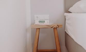 A wooden side table stands next to a bed with light bedding. A small lamp hangs on the wall above the table.