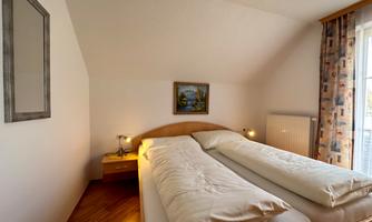A cozy bedroom with two single beds and a small painting on the wall. The room has large windows and warm wooden interior.