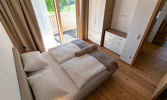 A bright bedroom with a double bed and large windows. The decor is modern and minimalist, with wooden flooring and neutral colors.