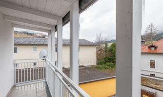 A balcony with a railing that offers a view of neighboring buildings and a green area. The sky is cloudy, and a tranquil environment is visible.