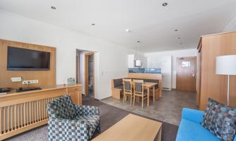 A modern living room with a blue couch and a patterned armchair. The room is bright and inviting, with a dining table and a kitchen corner.