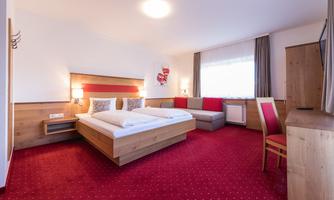 A bright hotel room with a double bed and a cozy seating area. The carpet is red and the walls are painted in neutral colors.