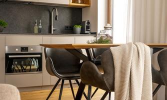 A modern kitchen with a wooden table and stylish chairs. A cozy scarf lies on the table and large windows provide plenty of light.