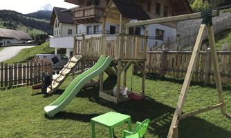A playground with a slide and a swing in a green garden. In the background, there is a beautiful wooden house.