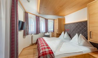 A cozy bedroom with wooden interiors and a large double bed. The windows are adorned with light curtains and a television is mounted on the wall.