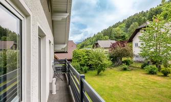 A balcony with a view of a green meadow and surrounding trees. In the background, some houses and hills can be seen.