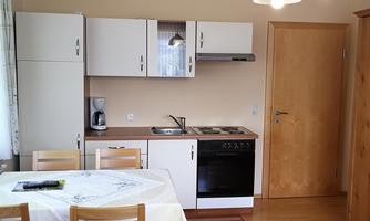 A modern kitchen with a dining table and wooden chairs. The walls are in soft colors, and there is a refrigerator as well as a cooking area.