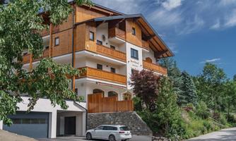 A modern house in Alpine style with wooden cladding and several balconies. In the foreground, there is a car, and it is surrounded by trees and greenery.