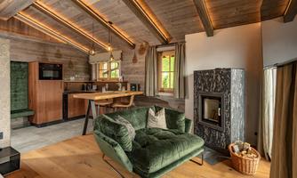A cozy living space with a wooden ceiling and modern furniture. In the background, a kitchenette and a fireplace can be seen.