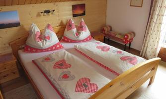 A cozy bedroom with a double bed and heart patterns on the bedcovers. The walls are made of wood, and there is a window with curtains.