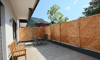 A beautiful terrace with wooden furniture and a privacy screen made of wooden boards. In the background, there are trees and a clear blue sky.