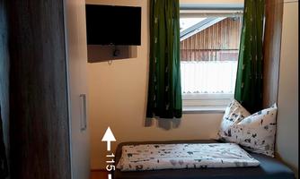 A cozy bedroom with two single beds, a TV, and green curtains. The walls are light, and there is a wooden wardrobe.
