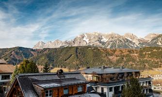 A picturesque mountain landscape with snow-capped peaks and a clear sky. In the foreground, traditional buildings and green forests can be seen.