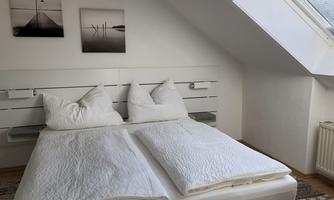 A bright bedroom with a double bed and two pillows. Three pictures hang on the wall, and there is a skylight that lets in daylight.