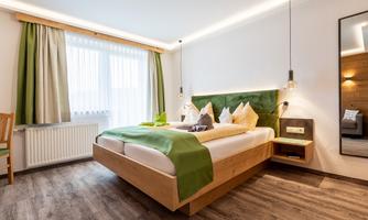 A modern bedroom with a wooden bed and green accents. Large windows provide plenty of daylight and a cozy atmosphere.