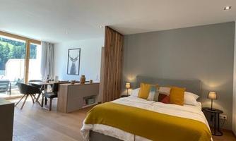 A modern bedroom with a large bed and colorful pillows. The room is bright and friendly with wooden flooring and a window that lets in daylight.