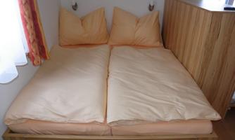 A spacious bed with orange bedcovers in a simple room. The walls are bright, and there is a wooden furniture arrangement.