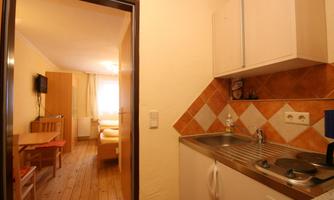 A cozy kitchenette with a sink and stove, located in a bright apartment. In the background, a dining area and a living area are visible.