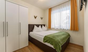 A cozy bedroom with a double bed and a large window. The walls are light, and a green throw is laid over the bedspread.