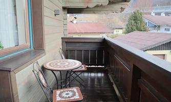 A cozy balcony with a small table and a chair. In the background, traditional houses and trees can be seen.