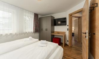 A modern room with two beds and light curtains. There is a desk, a television, and an open door to an adjacent room.