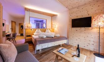 A stylish hotel room with wooden paneling and modern design. There is a double bed, comfortable seating, and a television.