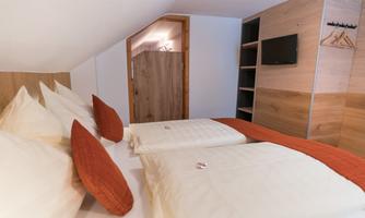 A cozy bedroom with two beds and pillows in warm colors. The room is modernly furnished and features a television and a wooden wardrobe.
