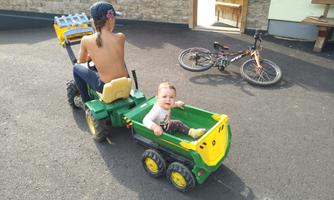 A child is riding a small tractor while a baby sits in the trailer and smiles. In the background, a bicycle is standing, and a wooden building is visible.