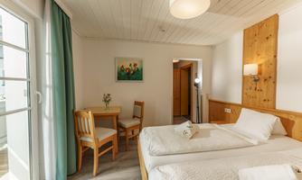 A cozy bedroom with two single beds, wooden furniture, and a small table. Large windows let in plenty of light and create an inviting atmosphere.