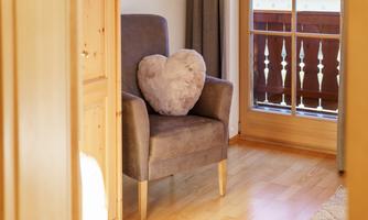 A comfortable armchair with a heart-shaped cushion is in a bright room. The windows offer a view of a balcony.