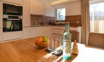 A modern kitchen with bright furniture and large windows. On the table sits a fruit bowl with apples and oranges, as well as a glass carafe.