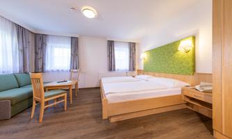 A bright hotel room with a double bed, a table, and two chairs. There is a comfortable couch and large windows that illuminate the room well.