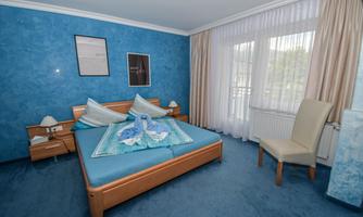 A modern bedroom with blue wall paint and carpet. The room features a large bed, a chair, and plenty of natural light through large windows.