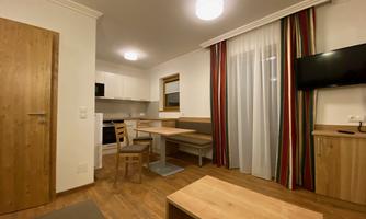 A modern living room with a small kitchenette and a dining table. The decor is bright and inviting, with wood and color accents.