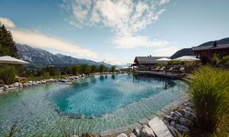 A relaxing outdoor pool surrounded by nature and mountains. The clear water surface reflects the blue sky and the clouds.