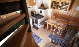 A cozy wooden room with a staircase, a dining table, and a cooking area. The space has warm colors and an inviting atmosphere.