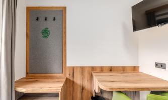 A modern living area with a wooden table and a green chair backrest. Some hooks are hanging on the wall, and a television is mounted.
