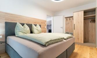 A modern bedroom with a large bed and green cushions. The room features a wardrobe and a bright, inviting atmosphere.