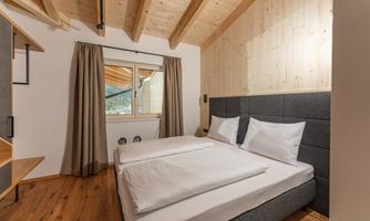 A modern bedroom with a double bed and bright wooden details. Large windows let in plenty of daylight and offer a view outside.